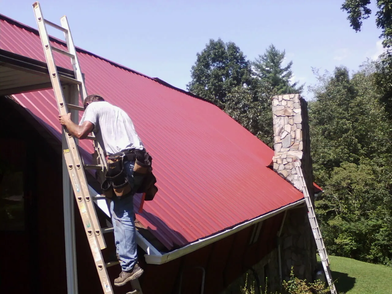 Roofer on a ladder beside a red metal roof and stone chimney — Soffit & Fascia Repair by Roofing Brothers in Thief River Falls